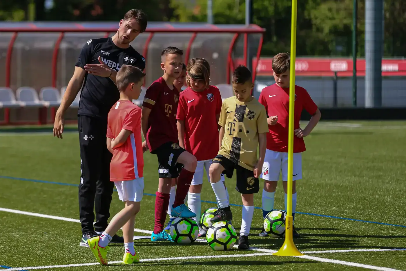 WePlay-trainer geeft instructies aan groep jeugdvoetballers met ballen op een zonnig kunstgrasveld