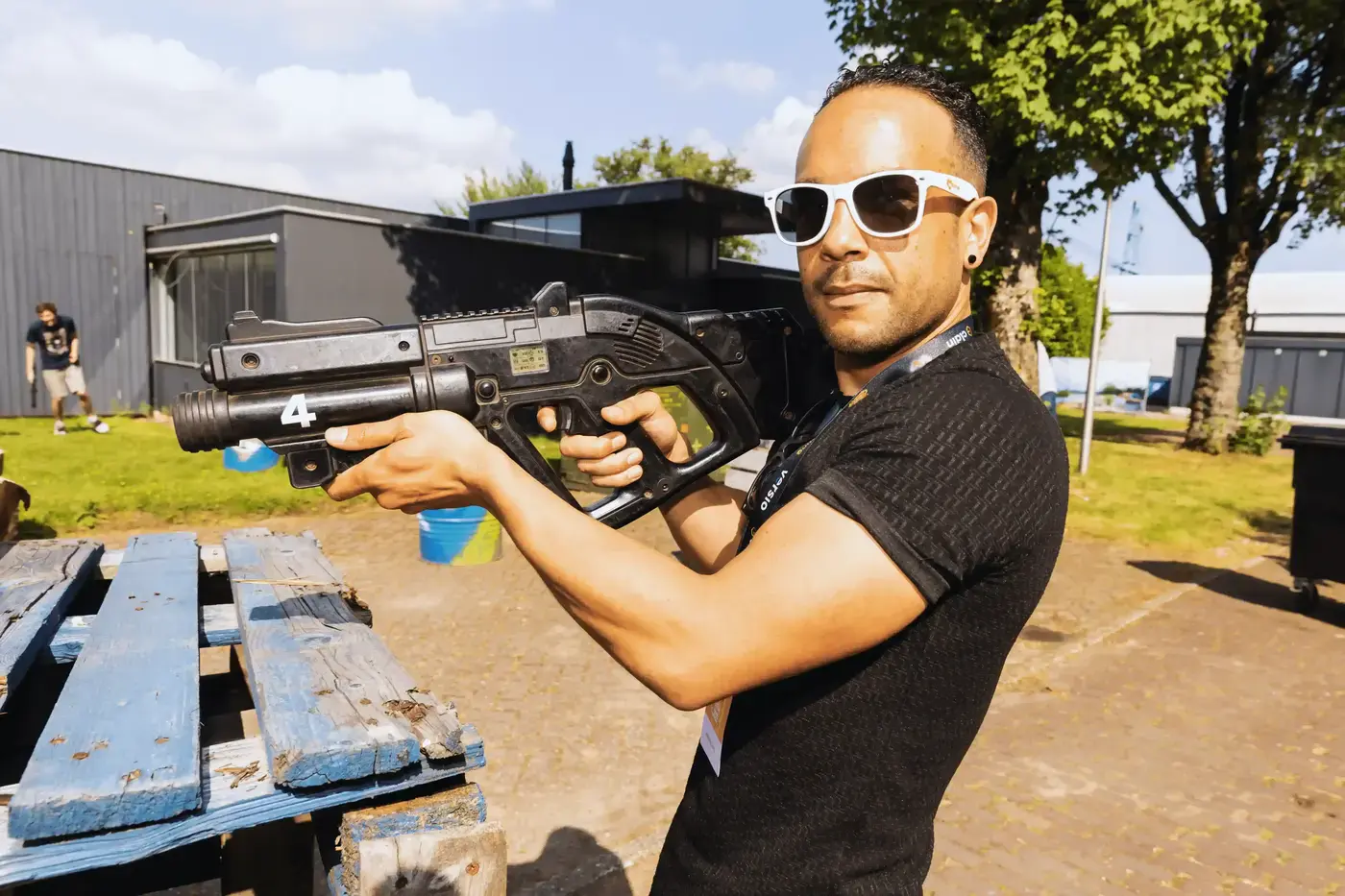 Man met zonnebril richt zijn laserwapen bij een picknicktafel tijdens een lasergame op een zonnig buitenterrein