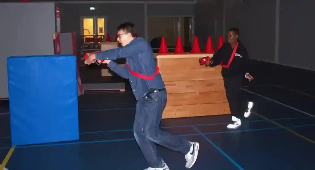 Twee spelers rennen met laserpistolen door een sporthal met obstakels van matten en pionnen